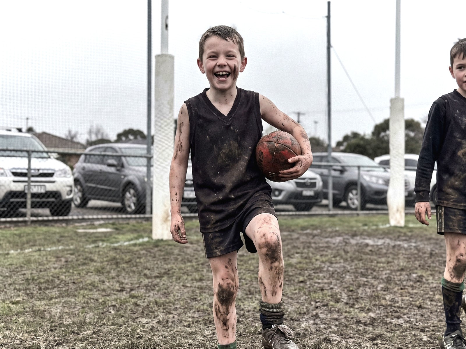 A muddy nine year old boy laughing after junior football, a ball under his arm